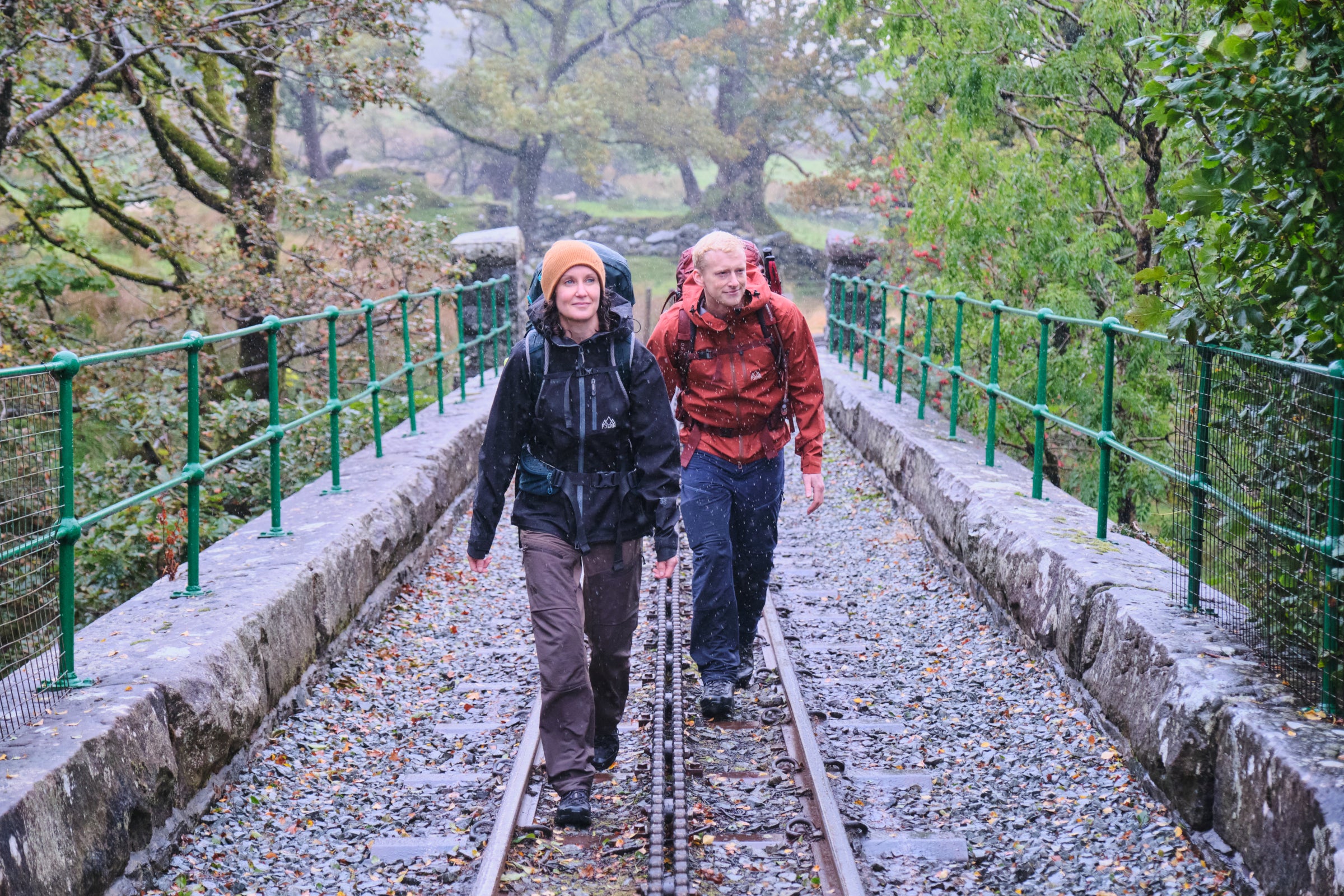 Two hikers in Fjern gear walk on a railway track in the rain