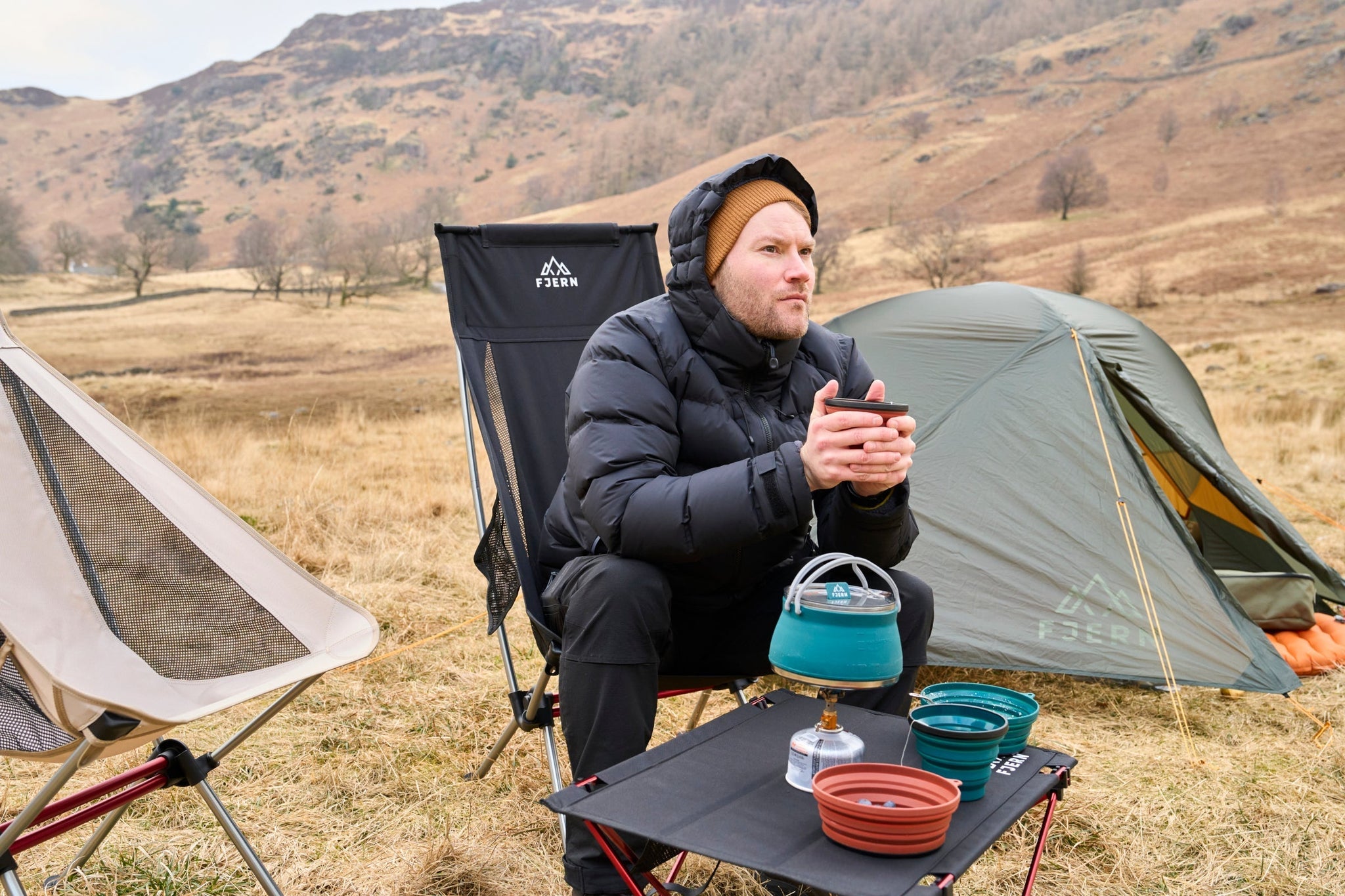 A man in a down jacket holding a mug at a campsite with Fjern camping equipment and tent