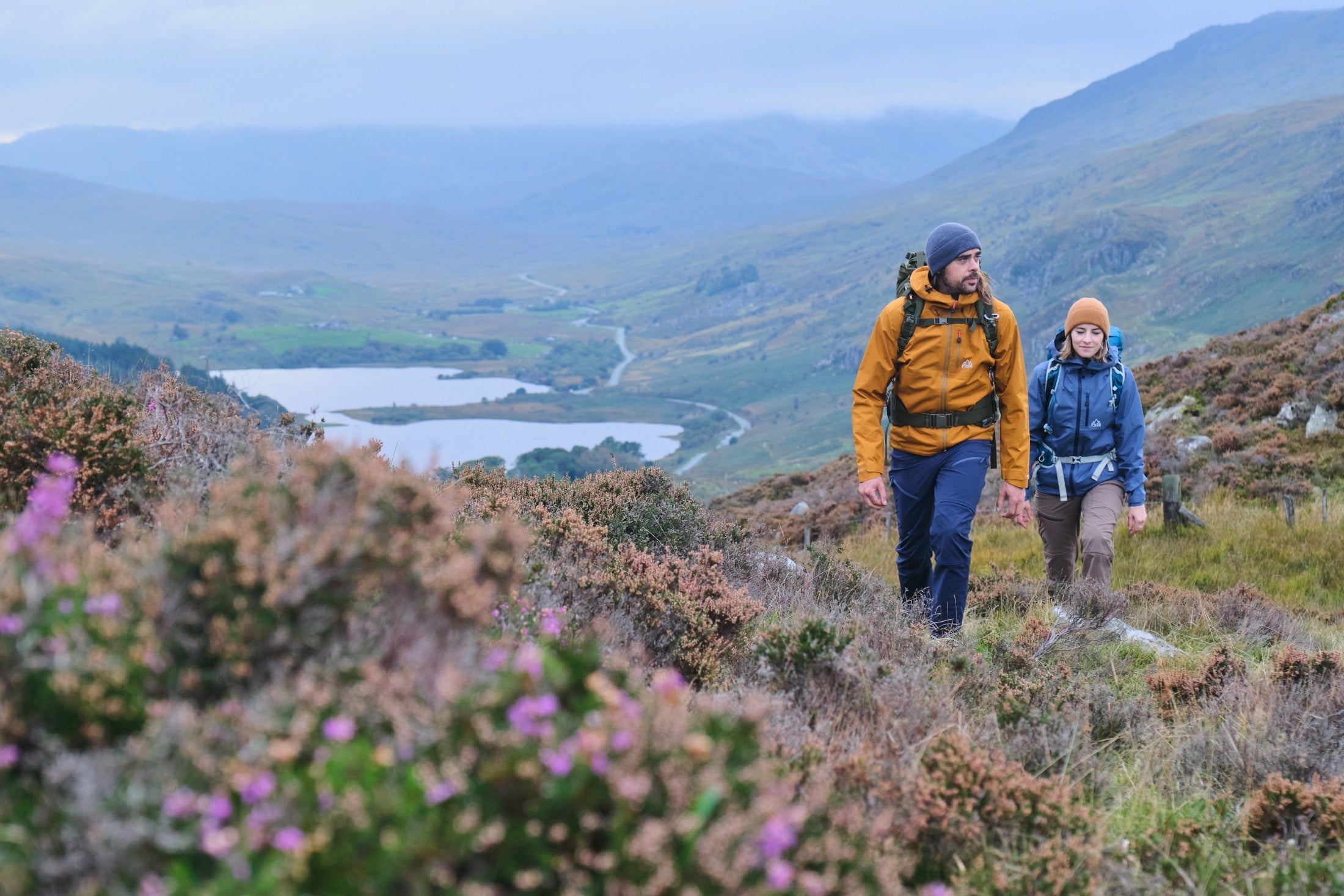 Hiking up a heather hillside above a distant lake in Forsvar Jackets