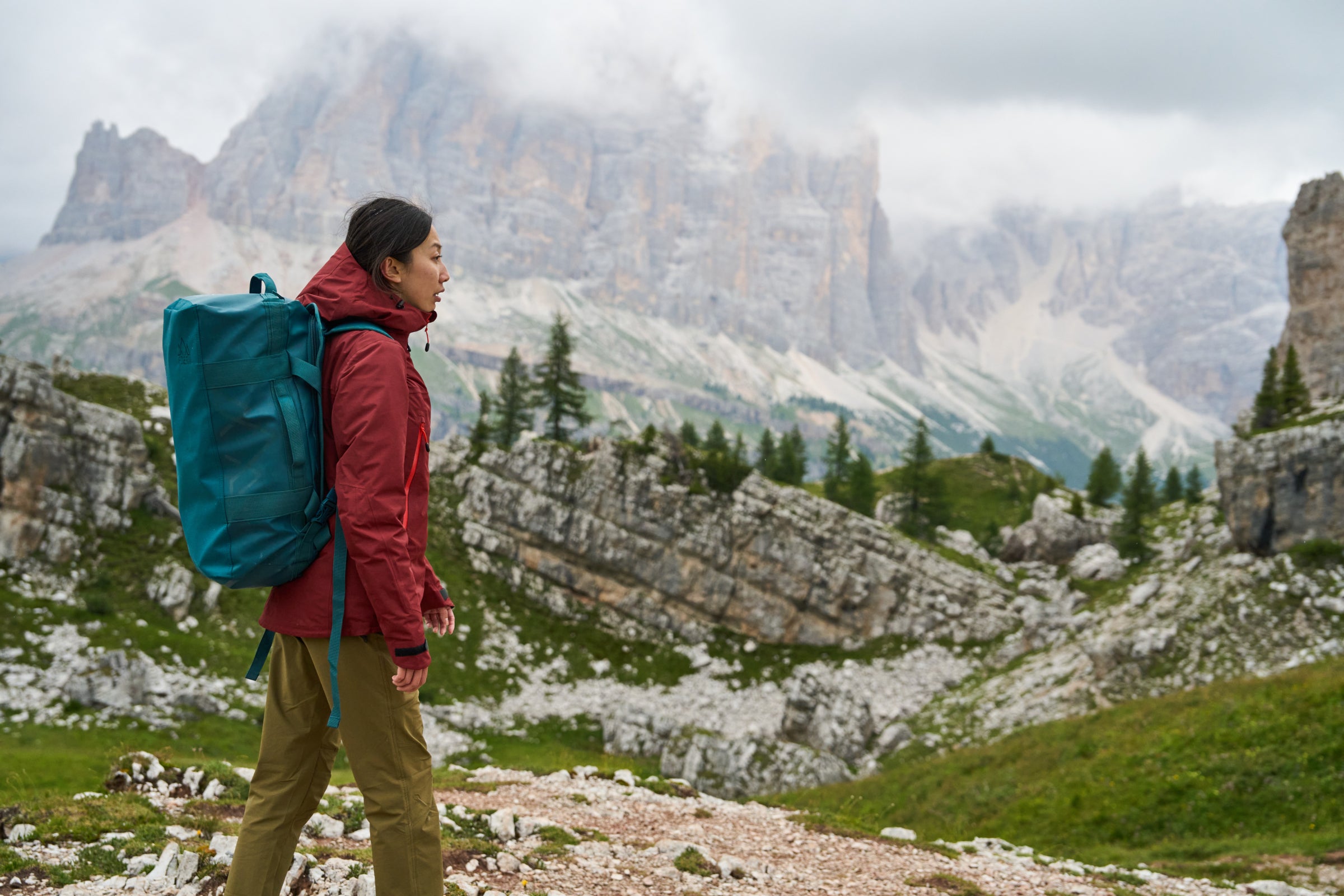 A woman wearing a Fjern jacket and carrying a large Fjern backpack gazes at a rugged mountain landscape, embodying a sense of adventure and exploration