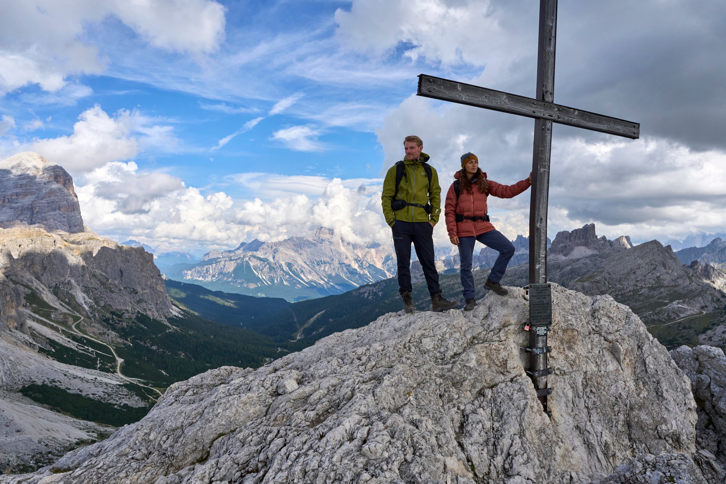 Two people standing on a rocky mountain peak beside a large wooden cross