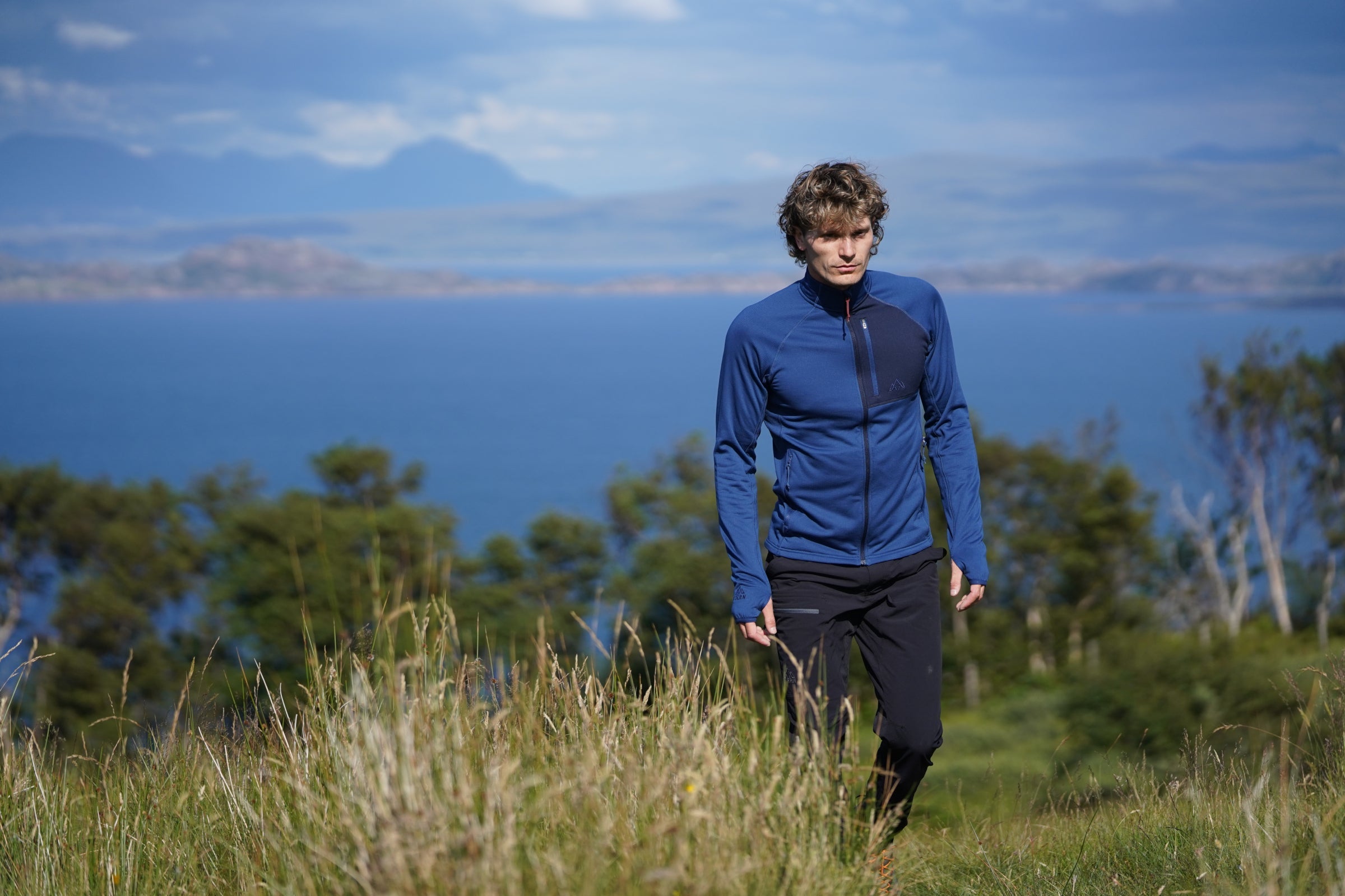 Man walking through grassy hillside in Fjern fleece jacket, overlooking the ocean