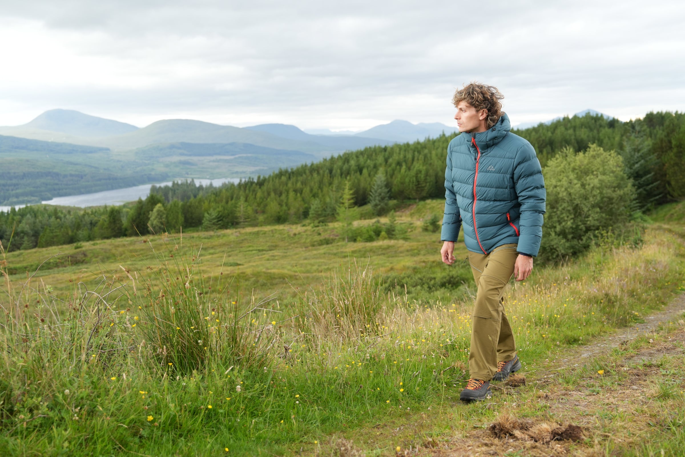 Man hiking in Fjern insulated jacket, walking along hillside with forest and mountains in the distance