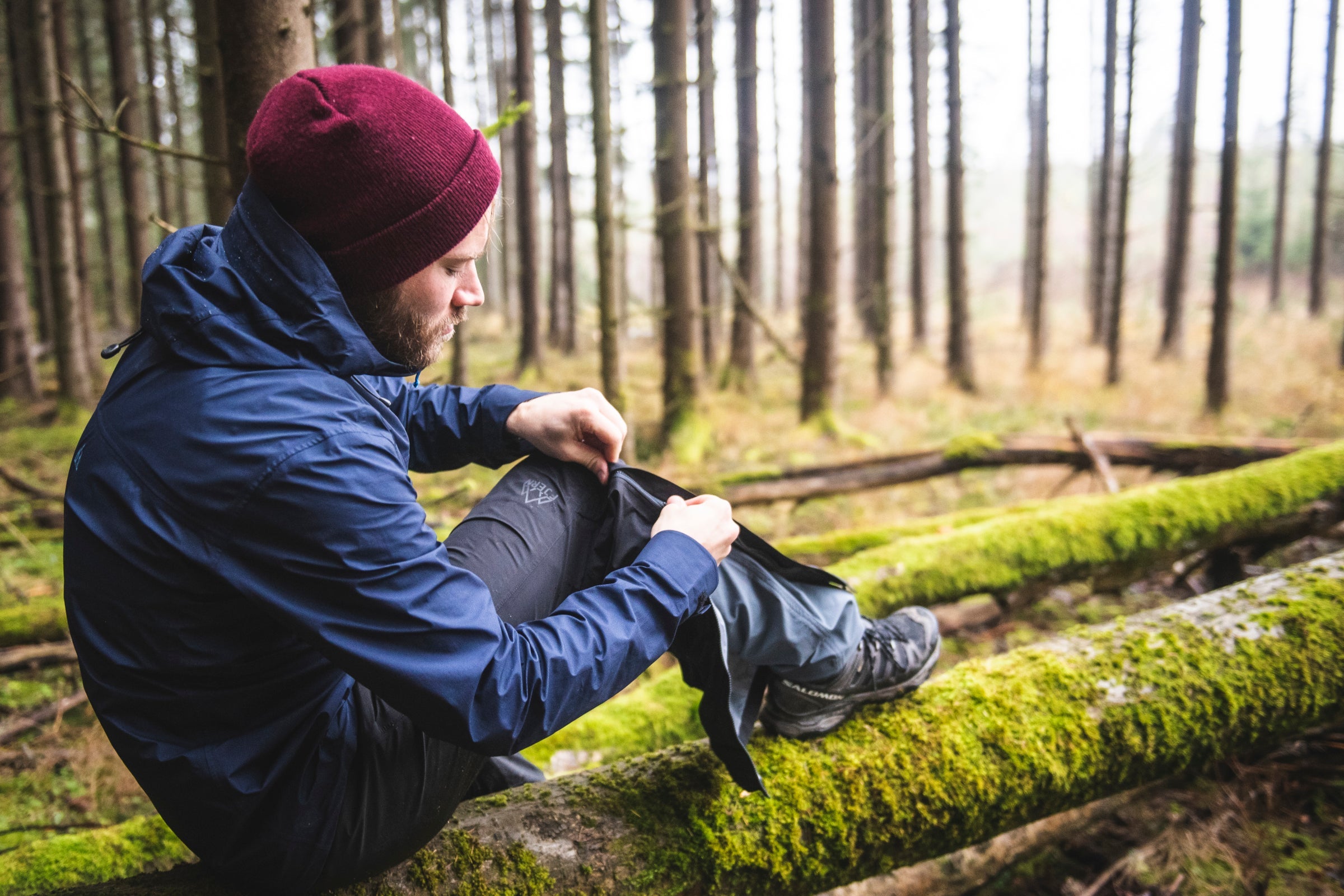 Man sitting on mossy log in forest adjusting Fjern waterproof trousers