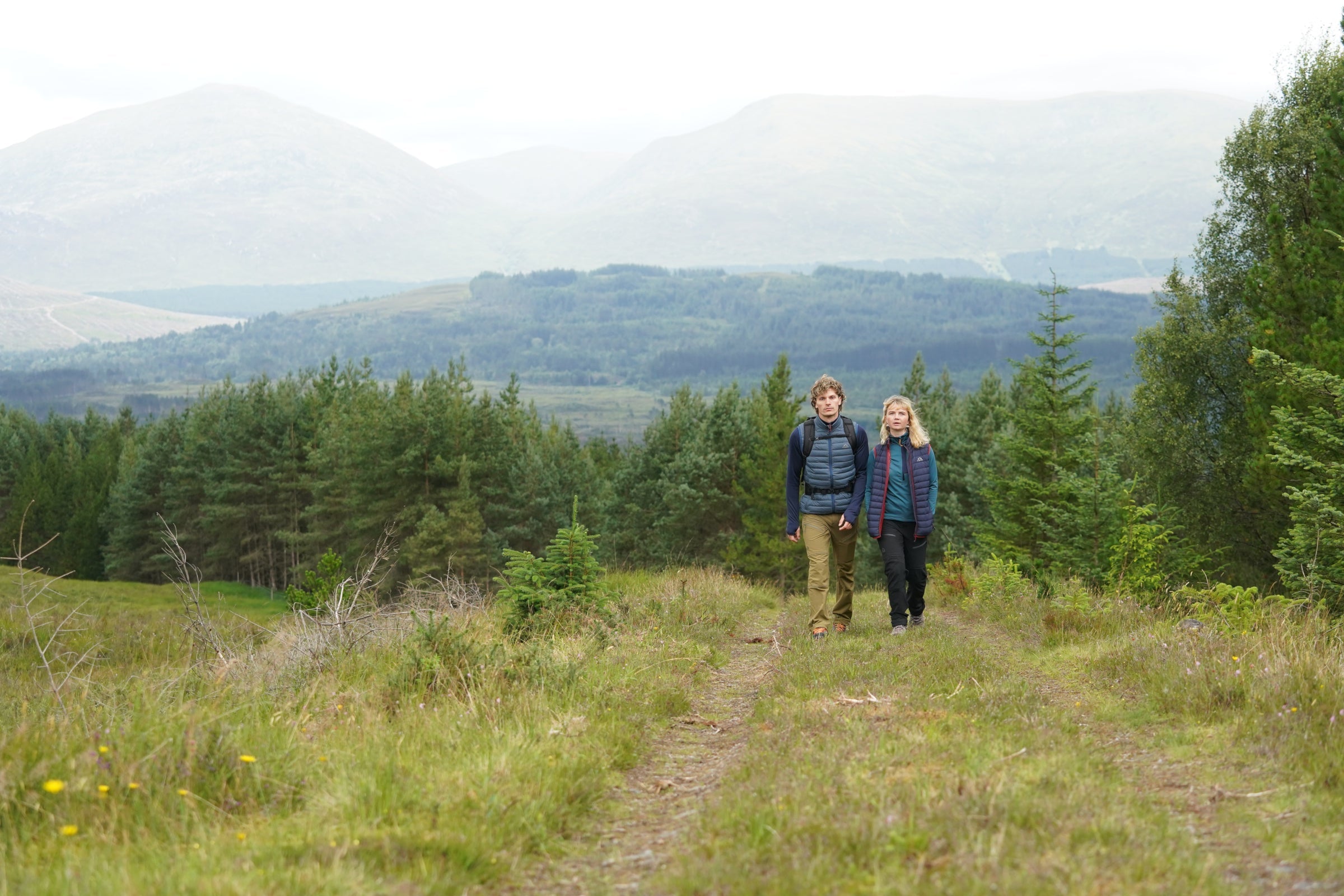 Two hikers wearing Fjern outdoor clothing walking on a forest trail with mountains in the background