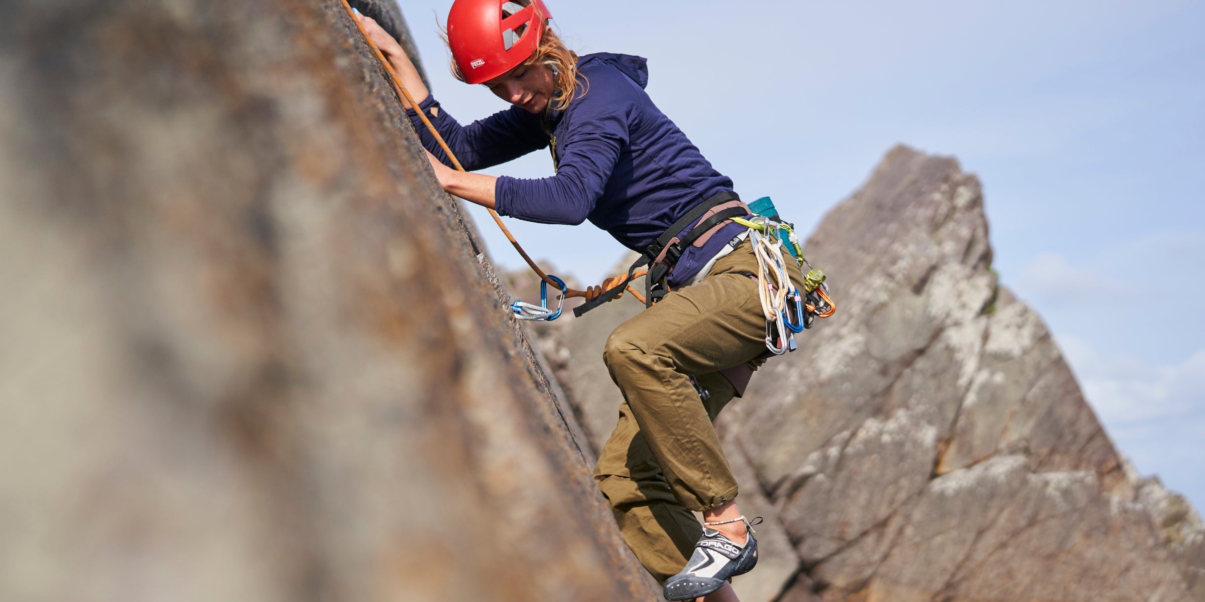 Woman climbing outdoors in Fjern climbing trousers, secured with harness and rope on coastal rock