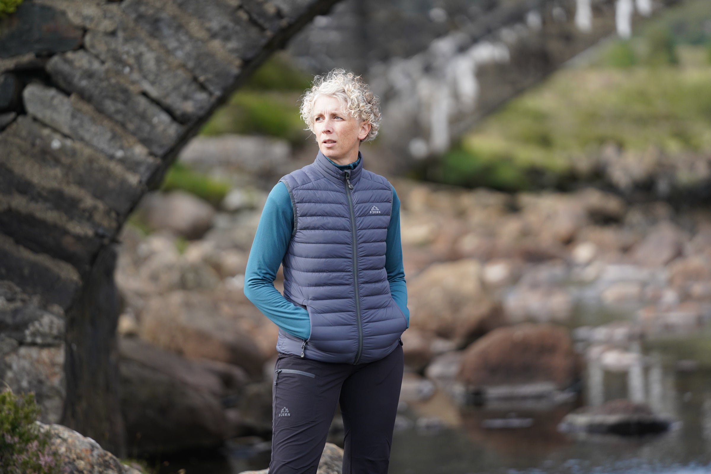 Woman wearing Fjern insulated gilet, standing outdoors by a stone bridge and river
