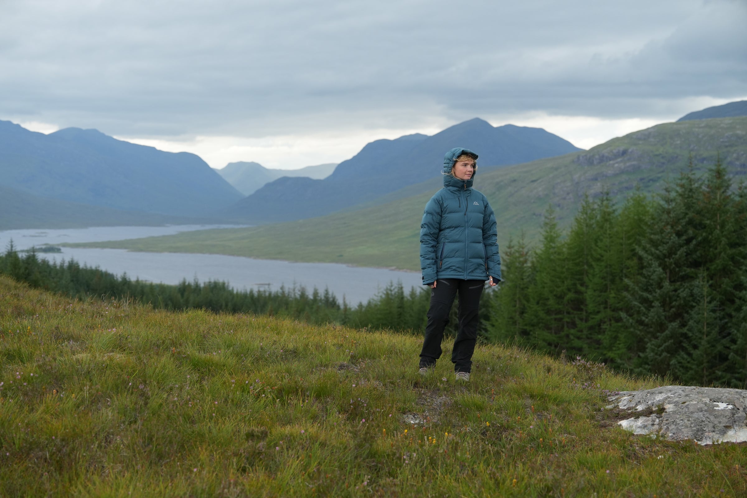 Woman standing outdoors in Fjern insulated jacket with hood, overlooking remote mountain and loch landscape