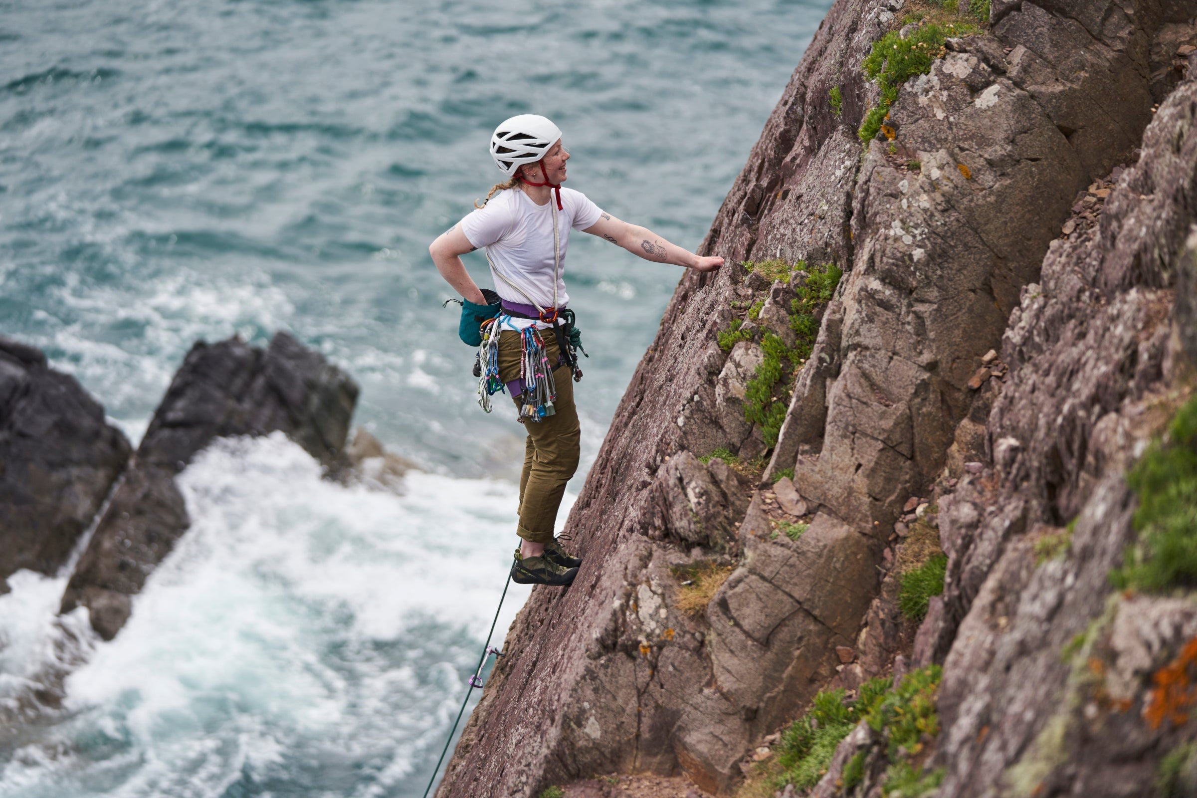 Woman rock climbing above the sea wearing Fjern short sleeve t-shirt and helmet