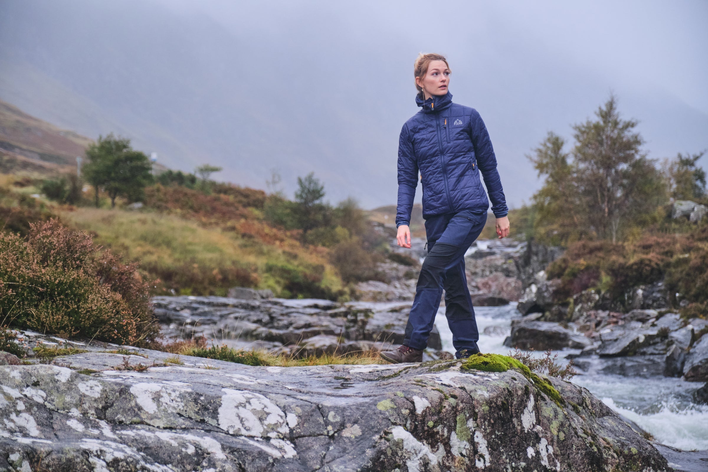 Woman hiking by a river in Fjern softshell trousers and insulated jacket