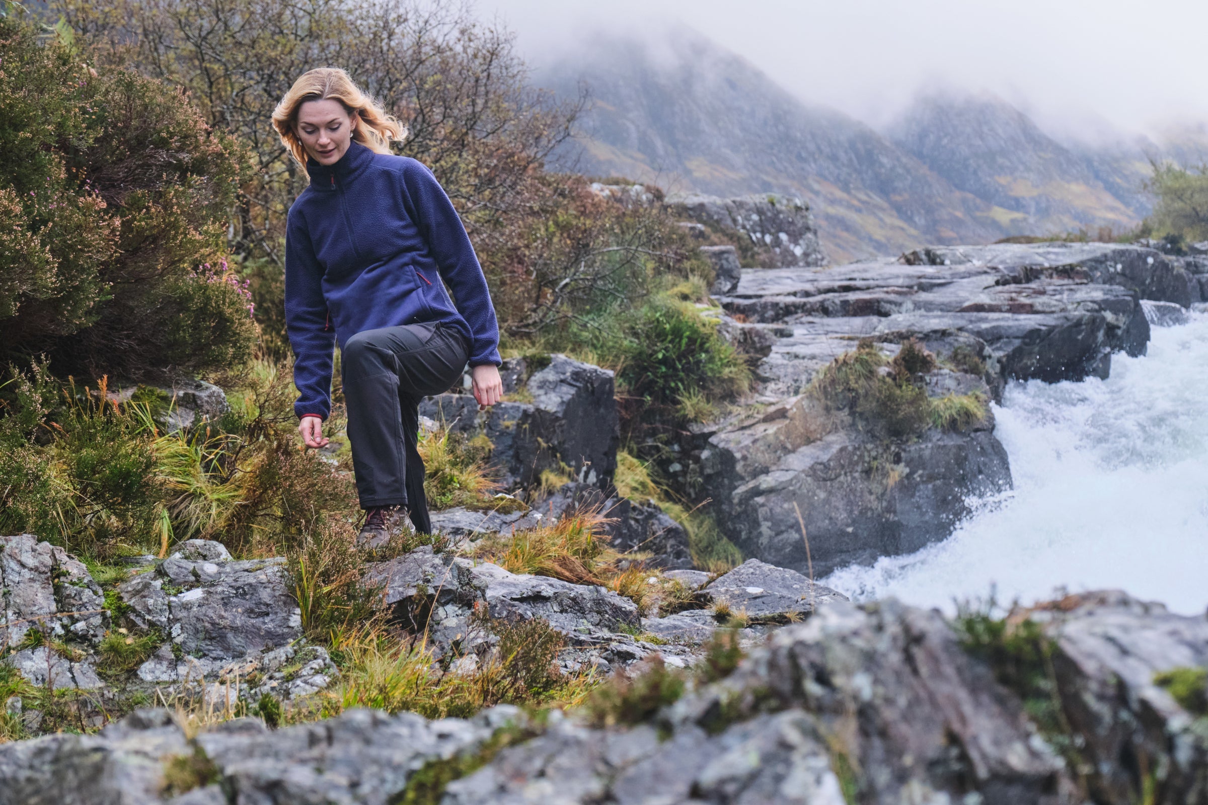 Woman climbing rocky trail beside waterfall in Fjern hiking trousers and fleece