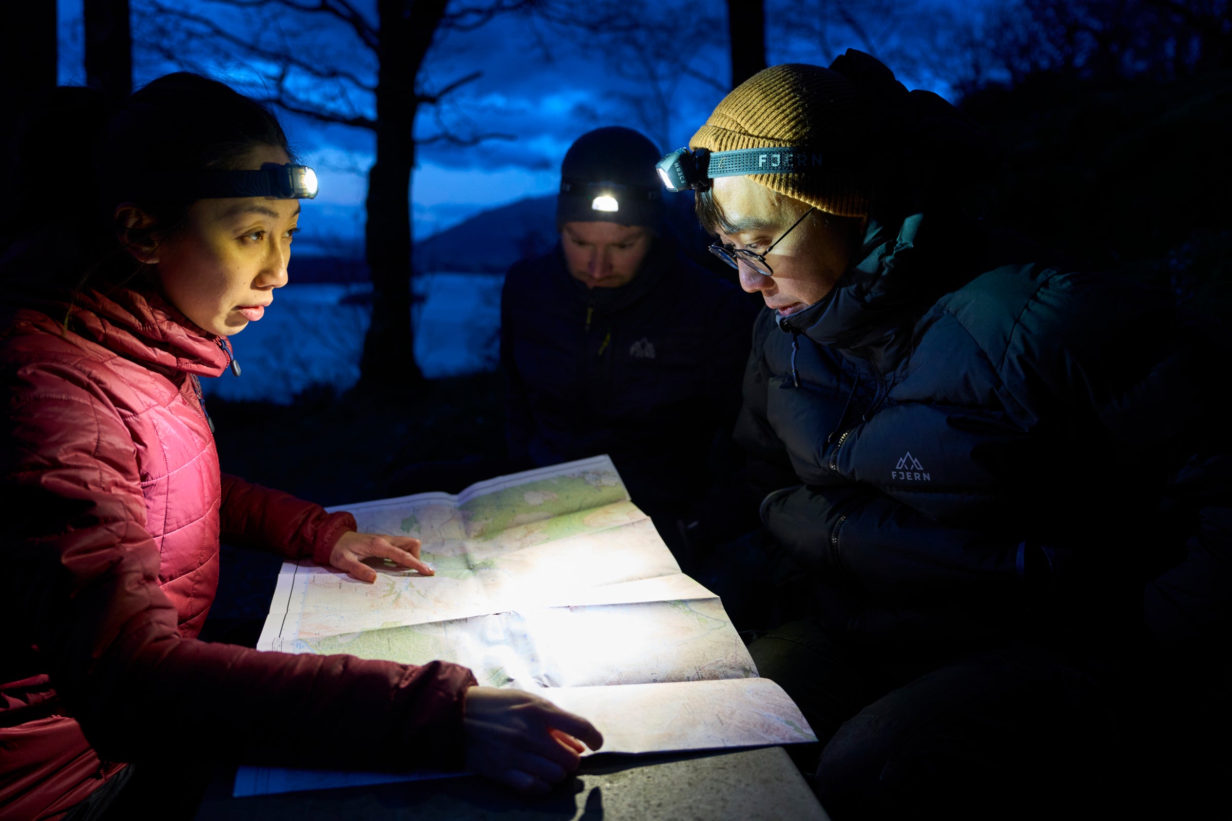Three people wearing headlamp and winter jackets sit together at night, studying a large map illuminated by their headlamps