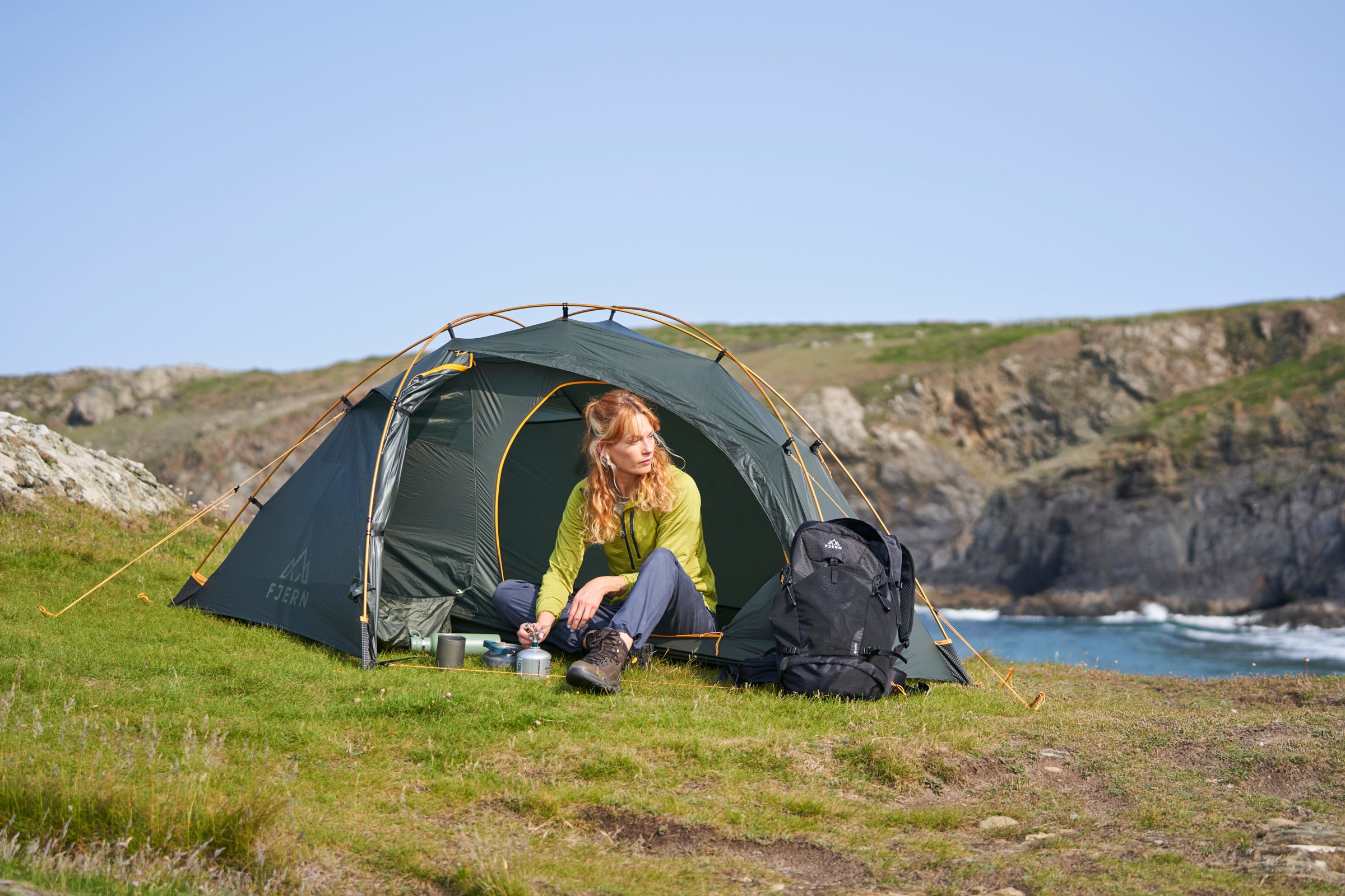 Camper sitting at Fjern tent entrance with cooking gear and coastal cliffs behind