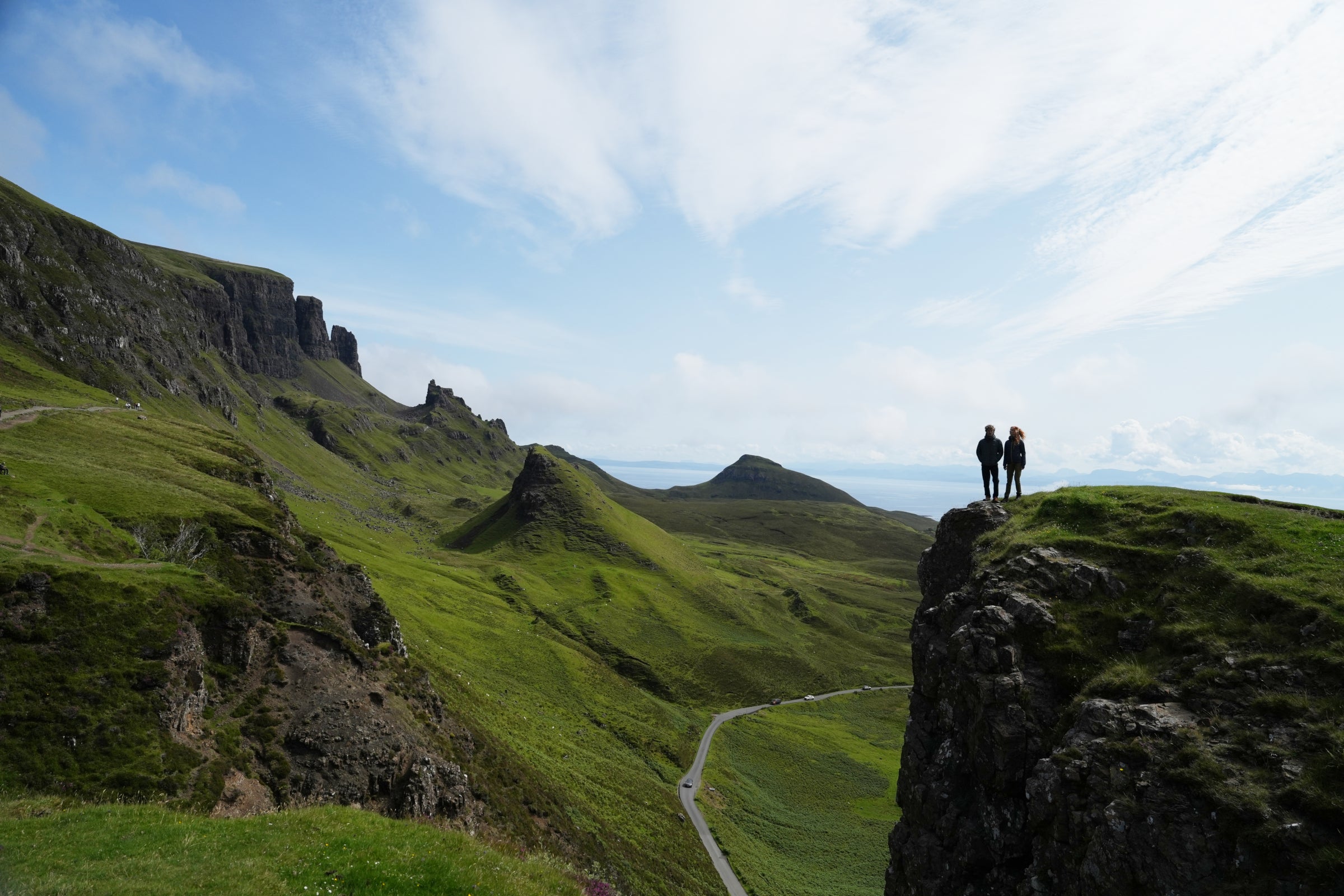 Two people standing on a rocky outcrop overlooking a scenic landscape with green hills and a clear sky