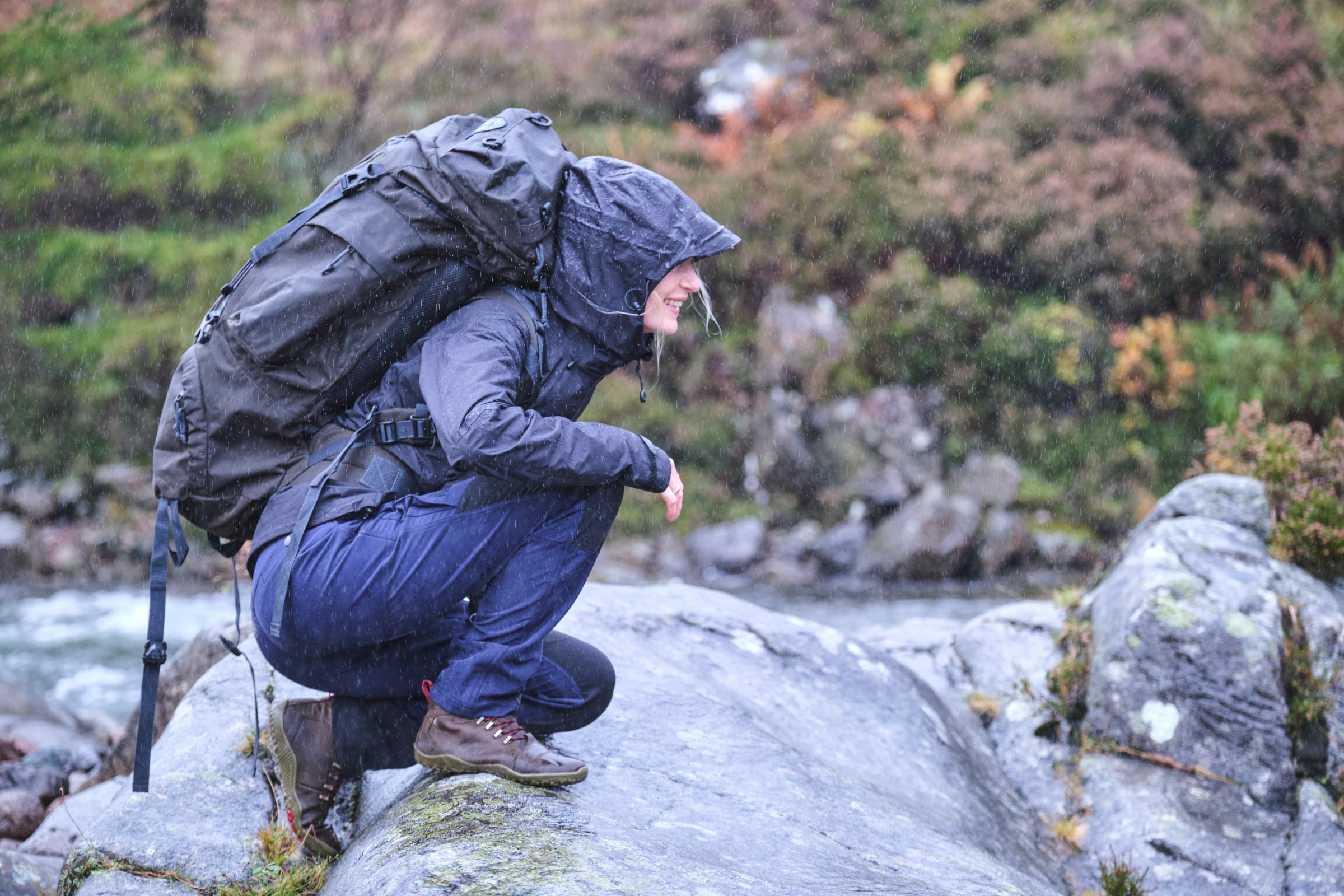 A hiker in Fjern clothing examines a stream on a rainy day