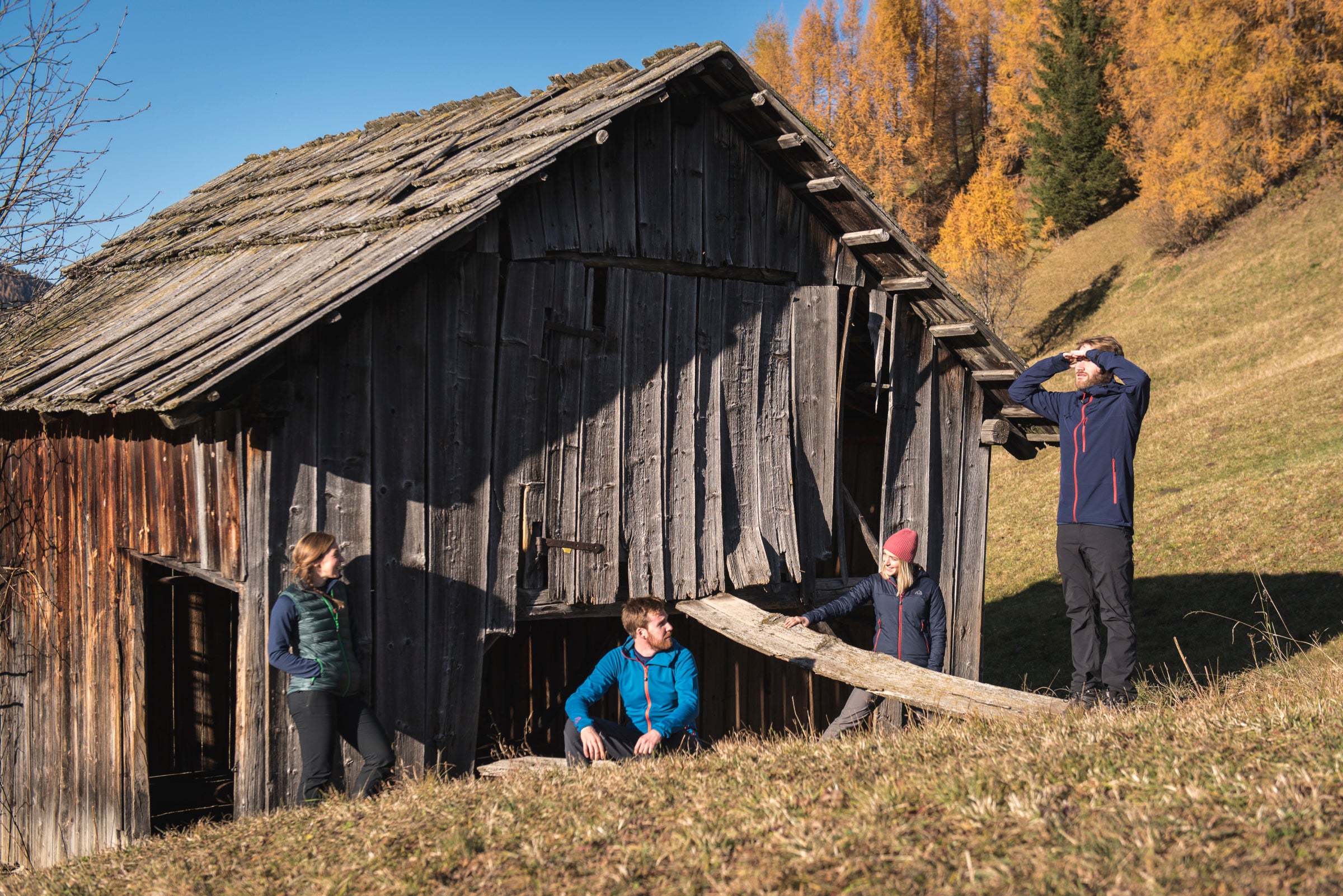 A group of four people wearing Fjern outdoor clothing explore and rest near an old wooden hut on a grassy hillside with autumn trees in the background