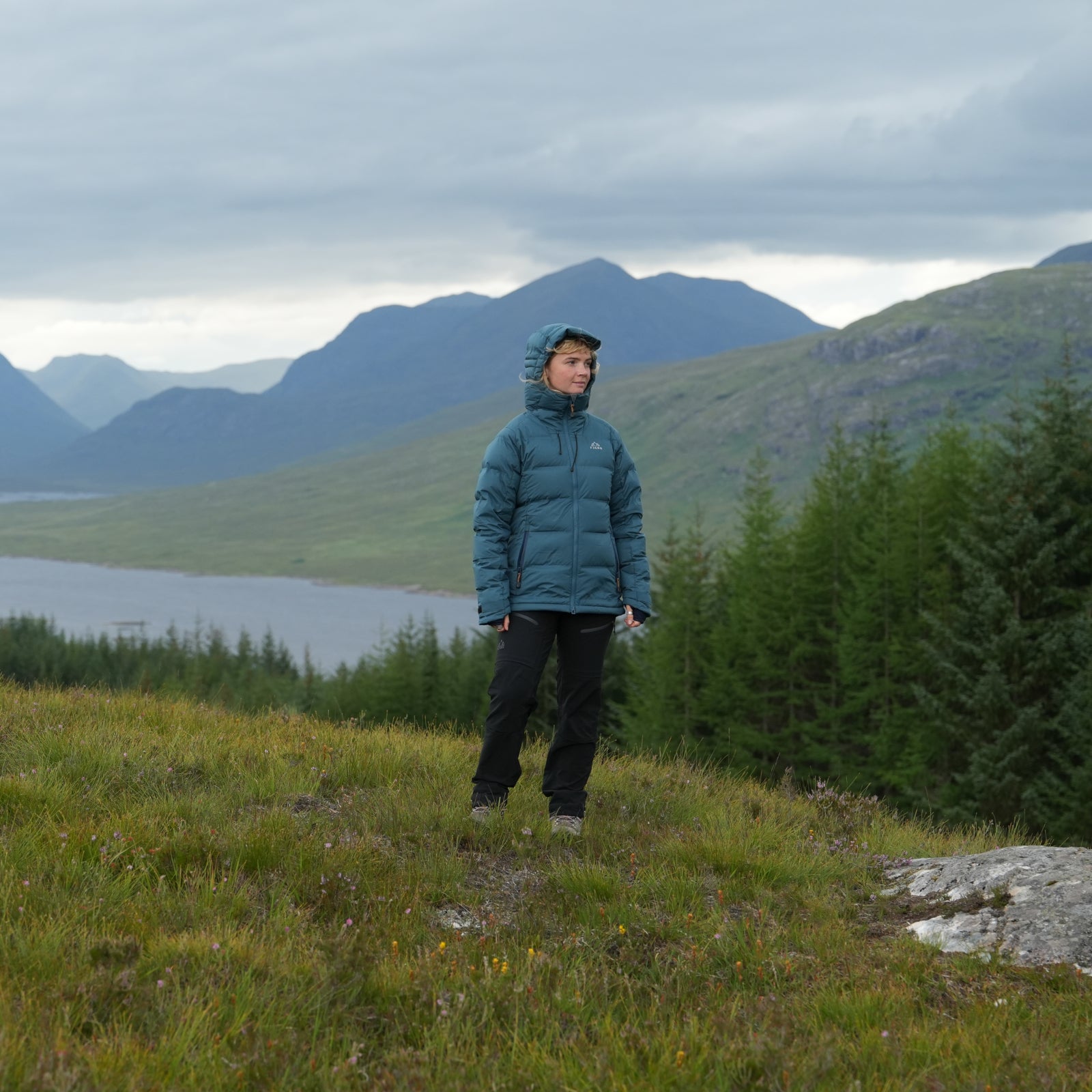 Woman standing outdoors in Fjern insulated jacket with hood, overlooking remote mountain and loch landscape
